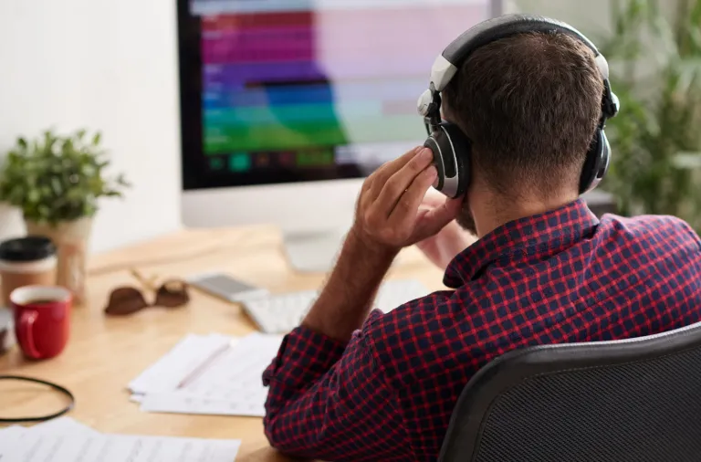 Mann mit Kopfhörern hört am Schreibtisch eine Aufnahme ab, auf dem Bildschirm farbige Audiospuren, daneben Notizblätter