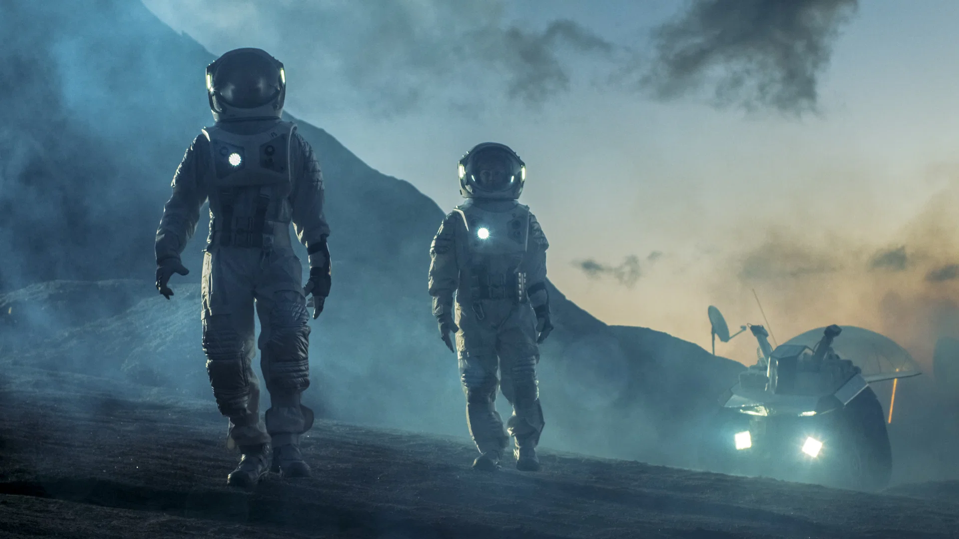 Zwei Astronauten in Raumanzügen gehen durch Nebel über felsigen Boden, rechts ein beleuchtetes Fahrzeug mit Antenne
