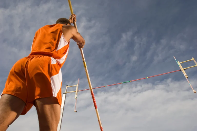 Stabhochspringer in orangem Trikot von unten gesehen, dahinter die Sprunglatte vor bewölktem Himmel