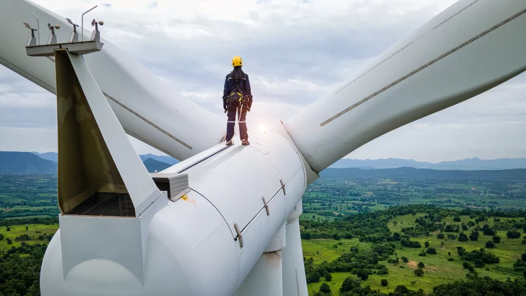 Techniker mit gelbem Helm steht auf der Gondel einer Windkraftanlage über grüner Landschaft