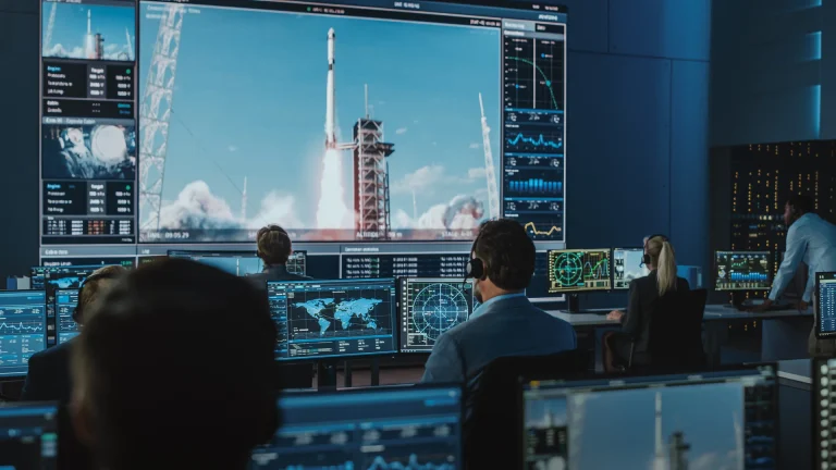 Mitarbeiter mit Headsets an Monitoren in einem Kontrollraum, auf der grossen Leinwand eine startbereite Rakete