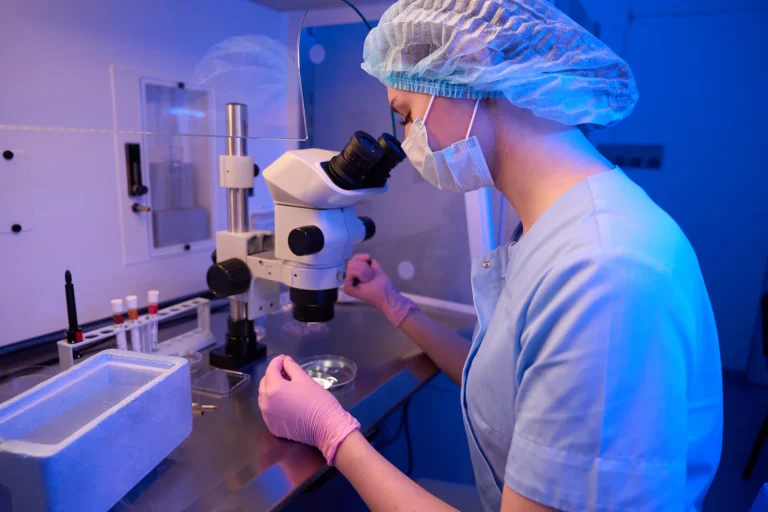 Laborant mit Haube und Maske am Stereomikroskop im blauen Licht